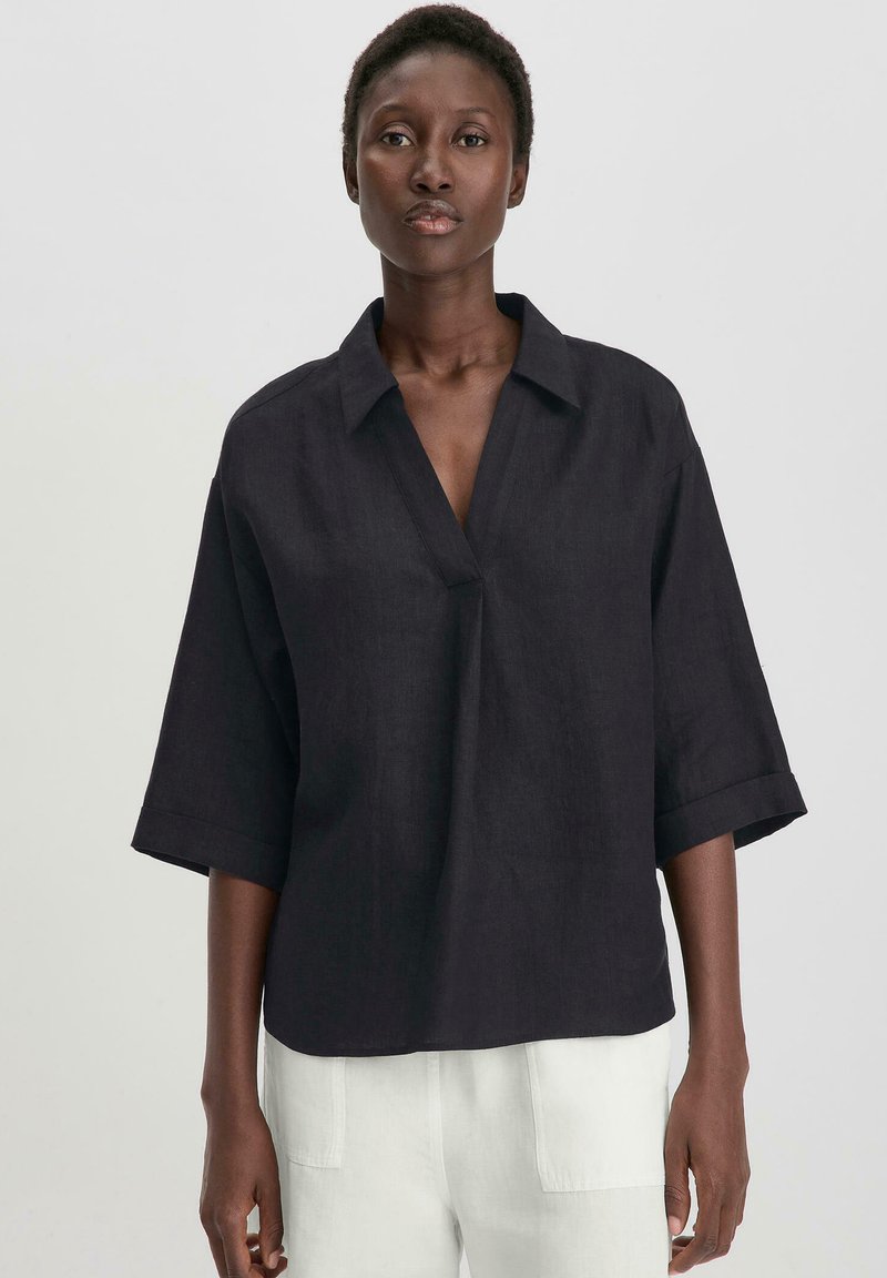Camicia nera in lino a maniche corte con collo a camicia e scollatura a V frontale. Il tessuto ha una texture sottile ed è di vestibilità rilassata.