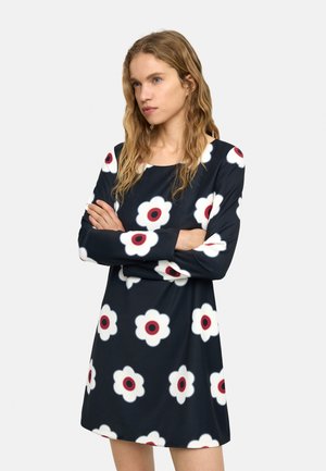 Kobieta o falowanych włosach w czarnej sukience z długim rękawem ozdobionej dużymi białymi i czerwonymi kwiatowymi wzorami, z założonymi rękami, patrząca na bok.