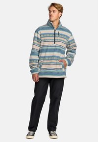 Joven con un pulóver de forro polar de patrón azul, blanco y beige, pantalones negros y zapatillas negras, de pie con una mano en el bolsillo.