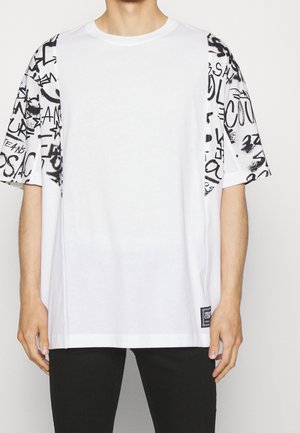 Biała oversize koszulka z czarnym tekstem w stylu graffiti na rękawach i bocznych panelach, noszona z czarnymi spodniami. Mała czarna metka w pobliżu dołu.