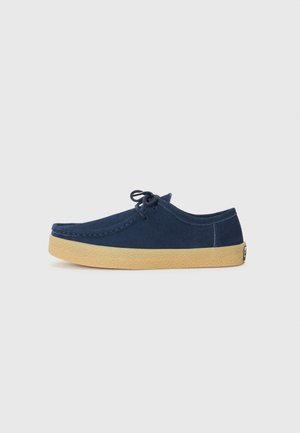 Zapato de ante azul oscuro con punta redonda, diseño con cordones y una suela de goma en color marrón claro con un acabado texturizado. Estilo minimalista, sin acentos adicionales.