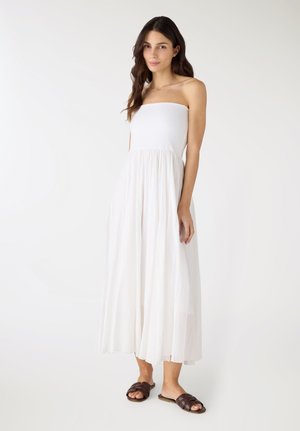 Donna in piedi con un vestito bianco senza spalline e sandali marroni senza lacci, su uno sfondo bianco semplice.