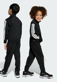 Černý teplákový komplet s mikinou na zip a kalhotami s elastickým pasem, které mají bílý trojproužek na rukávech. Kombinováno s černými sportovními botami.