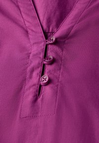 Lila Bluse mit V-Ausschnitt, vier passenden Knöpfen entlang der Knopfleiste und einem glatten, leichten Stoff mit subtiler Textur.