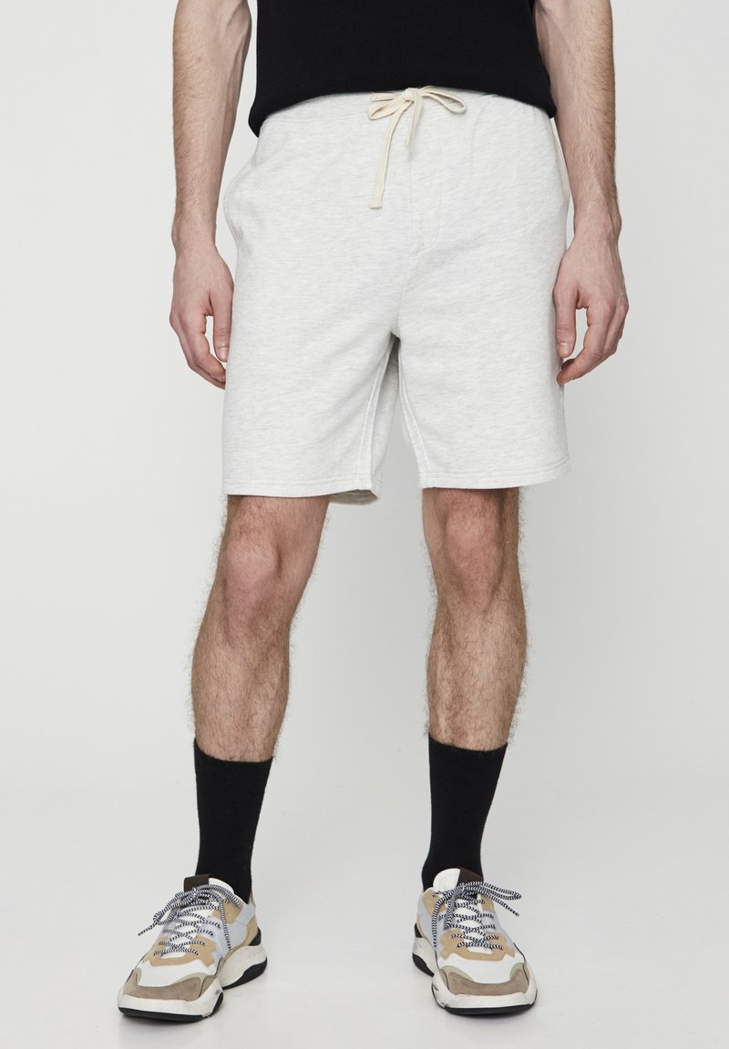 Shorts en coton gris clair avec une taille élastique et un cordon de serrage beige, comportant des coutures latérales. Le modèle porte des chaussettes noires et des chaussures multicolores.