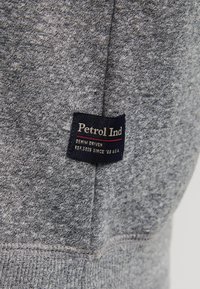 Tejido de sudadera gris con un acabado texturizado, que presenta una etiqueta que dice "Petrol Ind" en azul marino y blanco, junto con una línea de acento roja.