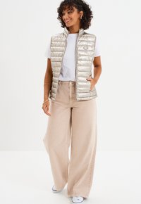 Metaliczna beżowa kamizelka puchowa z wysokim kołnierzem, z pionowymi przeszyciami i naszywką z logo, noszona na białej koszulce i beżowych spodniach o szerokich nogawkach.