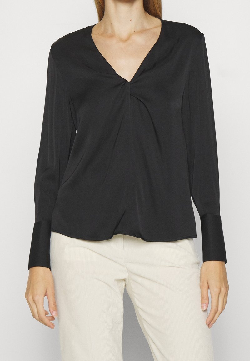 Maglietta nera a maniche lunghe con scollatura a V e dettaglio arricciato sul petto, realizzata in tessuto morbido; abbinata a pantaloni beige chiaro con texture.