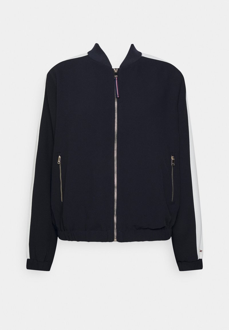 Blouson aviateur bleu marine avec des rayures blanches sur les manches, une fermeture éclair avant, deux poches zippées, et un col, des poignets et un ourlet côtelés.