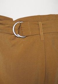 Cinturón de tela marrón con una hebilla circular de plata, que presenta una superficie texturizada y acentos cosidos. Pliegues visibles en la cintura.