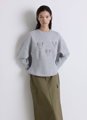 Frau mit langen dunklen Haaren trägt ein hellgraues Sweatshirt mit silbernen Kettendetails und einen olivgrünen Rock mit großen Taschen, steht vor einer weißen Wand.