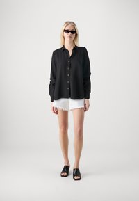 Camisa negra de botones con manga larga, que presenta un corte holgado. Combinada con pantalones cortos blancos deshilachados y sandalias negras deslizantes. Diseño minimalista.