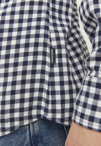 Camicia a scacchi blu navy e bianca realizzata in un tessuto dalla consistenza morbida. Presenta un piccolo etichetta del marchio sul lato della cucitura. I bordi sono rifiniti con cuciture ordinate.
