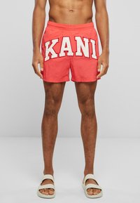 Red swim shorts with white "KANI" lettering. Elastic waistband, mid-thigh length, and smooth fabric. Worn with light-colored slide sandals.