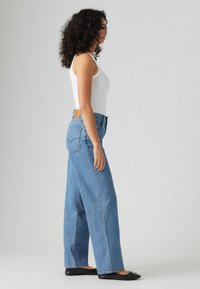 Hogewaisted, wijde blauwe spijkerbroek van denim, met een patch op de achterkant en stiksels details. Gecombineerd met een aansluitend wit tanktop.
