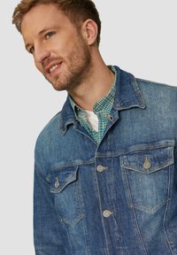 Jeansjacke in mittlerem Blau mit kontrastierendem Nähen, zwei Brusttaschen mit metallischen Knöpfen und einem klassischen Kragen über einem grün karierten Hemd.