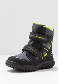 Botte d’hiver enfant noire et grise avec des accents jaune néon, deux sangles en velcro et une semelle à crampons robuste.