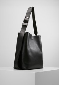 Czarna skórzana torba typu tote o miękkiej fakturze, z regulowanym paskiem i srebrnym zapięciem. Charakteryzuje się minimalistycznym designem i subtelnym brandingiem.
