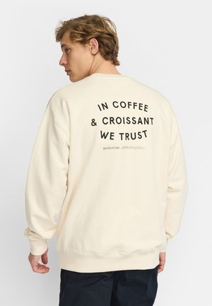 Jonge man met licht haar die een beige sweatshirt draagt met de tekst "IN COFFEE & CROISSANT WE TRUST" op de rug, staand tegen een eenvoudige achtergrond.