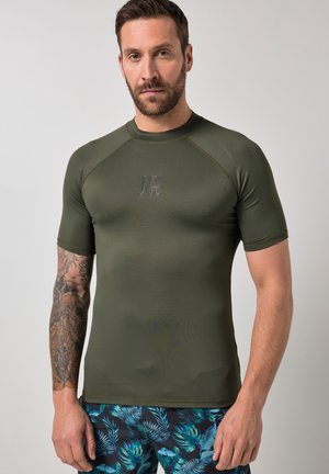 Homme avec barbe et tatouage sur le bras gauche, portant une chemise ajustée à manches courtes de couleur vert olive et un short bleu à imprimé tropical, sur un fond uni.
