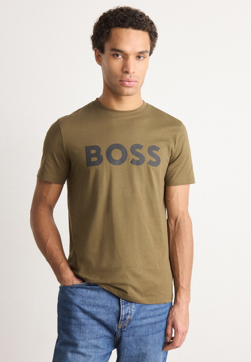 Alyvuogių žalios spalvos trumpomis rankovėmis marškinėliai su dideliu juodu "BOSS" logotipu priekyje. Minkšta medžiaga ir klasikinis apvalus iškirpimas. Dėvimi su mėlynomis džinsinėmis kelnėmis.