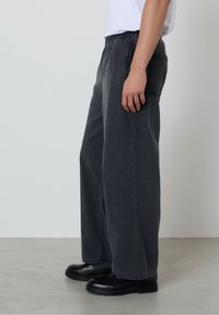 Oseba, oblečena v široke temnosive hlače, črne vezalke in belo majico s kratkimi rokavi, stoji na gladkem sivi tla.