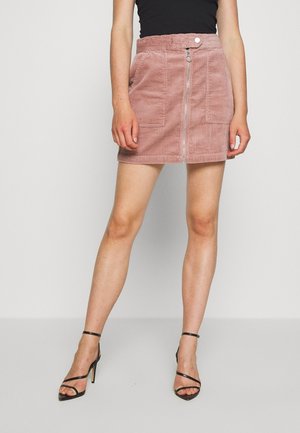 Kvinde iført en lyserød mininederdel i fløjl med lynlås foran og lommer, sort top og sorte høje hæle med ankelremme.