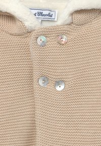 Beige stickad plagg med en mjuk krämfärgad fuskpälskrage, prydd med tre runda, glänsande knappar med ingraverade mönster framtill.