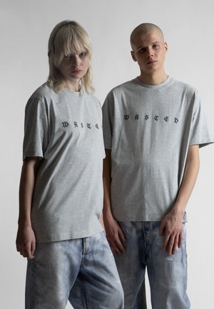 To unge voksne iført grå t-skjorter med gotisk tekst, den ene med teksten "WHITE" og den andre "WASTED," kombinert med løstsittende blå jeans.