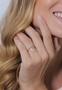 Goldring mit zwei strukturierten Ginkgo-Blatt-Akzenten, verstellbarer Band, getragen an einer Hand mit langen, lockigen blonden Haaren im Hintergrund.