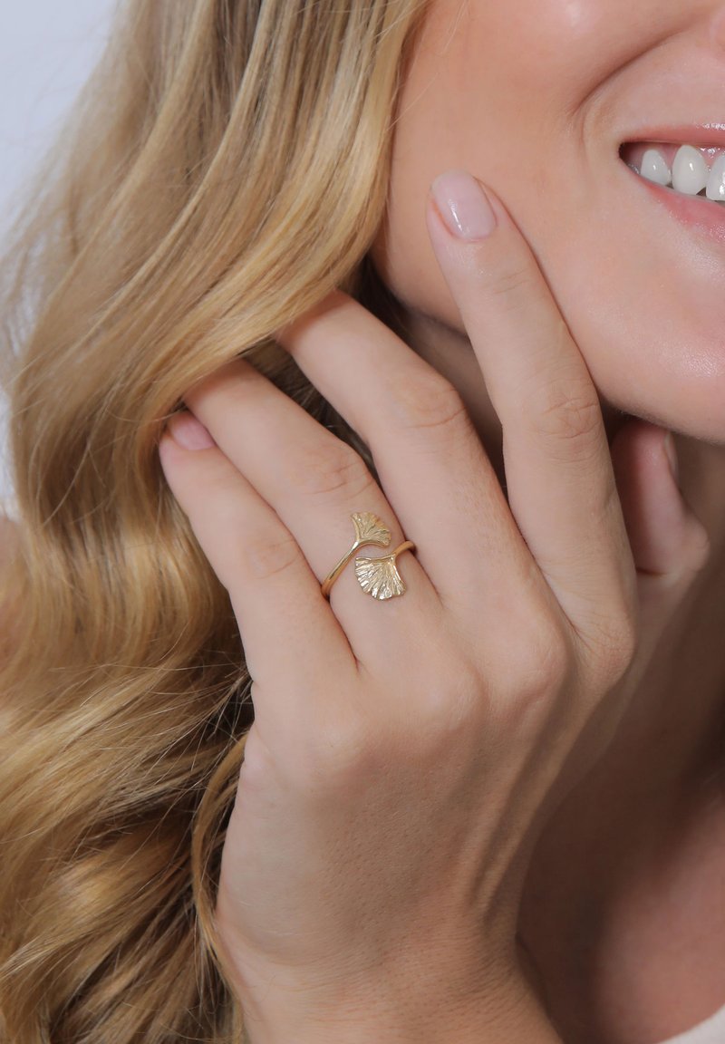 Goldring mit zwei strukturierten Ginkgo-Blatt-Akzenten, verstellbarer Band, getragen an einer Hand mit langen, lockigen blonden Haaren im Hintergrund.