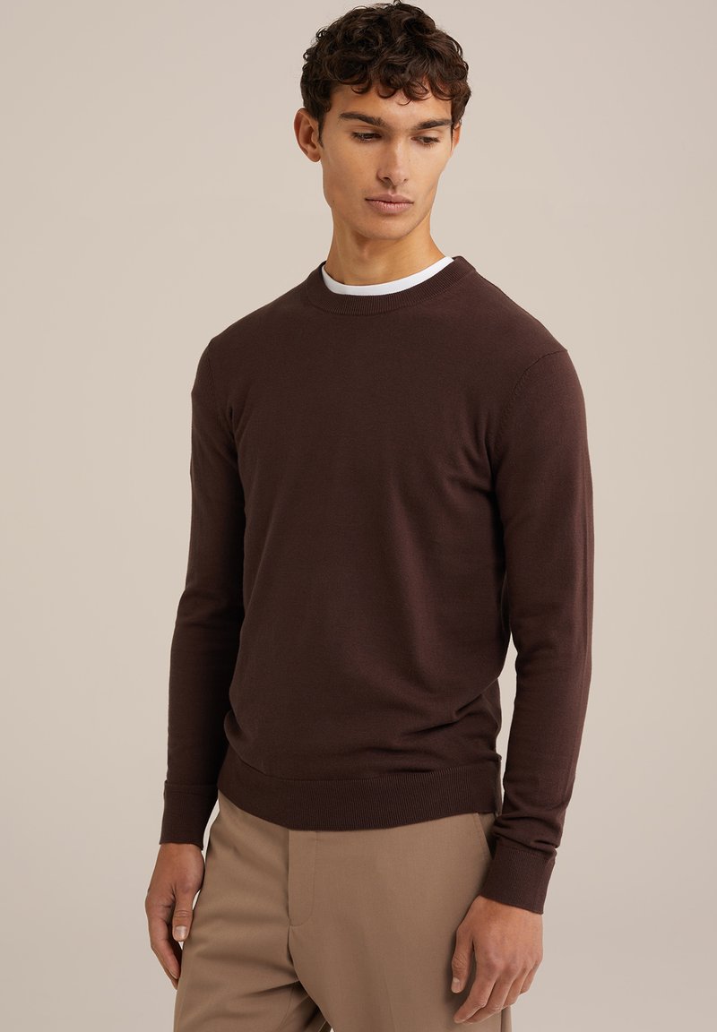 Suéter marrón de punto con cuello redondo, puños y dobladillo acanalados, hecho de una tela suave. Llevado sobre una camiseta blanca, con un diseño ajustado.