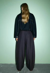 Übergroßes, dunkles Denim-Hemd mit sichtbaren Nähten, kombiniert mit lockeren, dunklen Hosen mit einer Gesäßtasche. Modell steht mit dem Rücken zur Kamera vor einer gedeckten grünen Wand.