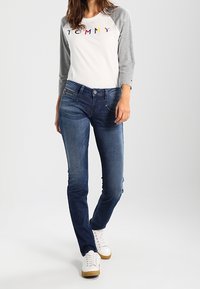 Weißes und graues Raglan-Shirt mit "TOMMY"-Aufschrift, kombiniert mit dunkelblauen Jeans. Weiße Sneaker mit Kautschuksohlen. Einfaches Design, lässige Passform.
