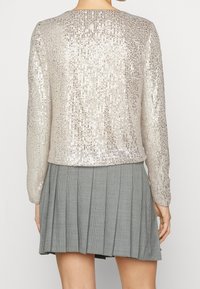 Béžový top s flitry, dlouhými rukávy, kulatým výstřihem a rovním lemem, zkombinovaný s šedou plisovanou sukní s kostkovaným vzorem.