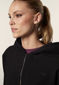 Hoodie zippé noir en tissu doux, doté d'une capuche, d'une fermeture éclair sur le devant et d'un petit logo brodé sur la poitrine gauche.