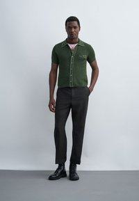 Mannequin masculin portant une chemise en maille verte à manches courtes, un pantalon noir, des bottes de combat noires et des colliers superposés, debout devant un fond gris clair.