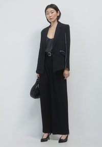 Donna che indossa un blazer nero su misura, pantaloni a gamba larga, top in satin e scarpe slingback con tacco, mentre tiene in mano una borsa nera su uno sfondo chiaro.