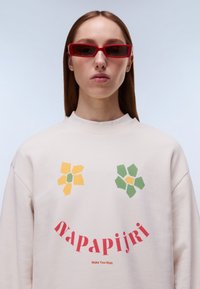 Μπεζ φούτερ με λουλουδένια σχέδια σε κίτρινο και πράσινο χρώμα· κόκκινο κείμενο λογότυπου στο κάτω μέρος. Το μοντέλο φοράει κόκκινα ορθογώνια γυαλιά ηλίου.