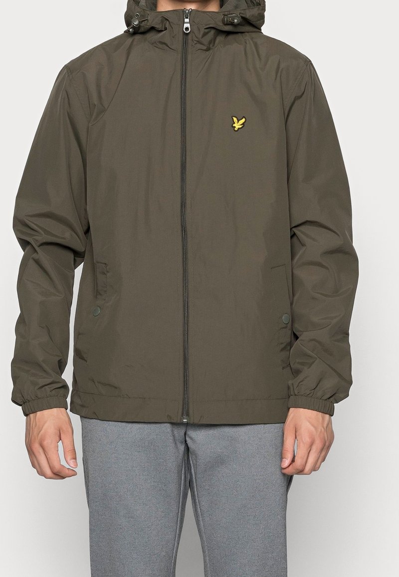 Homme portant une veste à capuche zippée vert olive avec des poignets élastiques et un petit logo d'oiseau jaune sur la poitrine gauche, assortie à un pantalon gris.