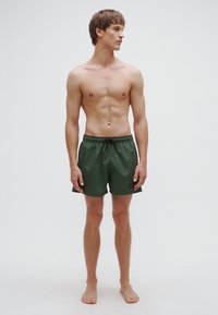 Zaļi peldbikses ar elastīgu jostasvietu un auklu. Izgatavotas no viegla materiāla, ar logo uz kreisās kājas. Modelis stāv basām kājām.