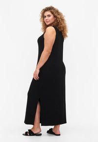 Černé, bezramenné maxi šaty se bočním rozparkem, vyrobené z měkké látky. Modelka má na sobě černé sandály s volánkovými detaily.