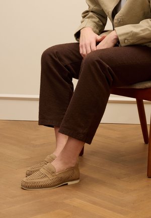 Tan loafers met geweven details, platte zool en gevoerde voetbedden, gecombineerd met bruine, gestructureerde, cropped broeken. Houten vloer op de achtergrond.