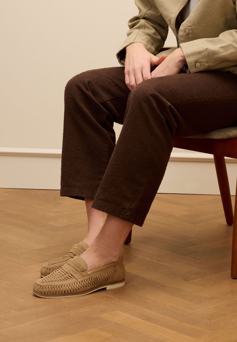 Tan loafers met geweven details, platte zool en gevoerde voetbedden, gecombineerd met bruine, gestructureerde, cropped broeken. Houten vloer op de achtergrond.