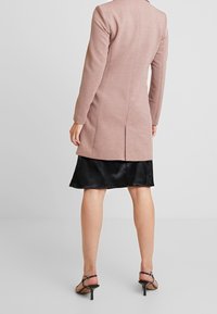 Jasnoróżowy płaszcz o dopasowanym kroju, z długimi rękawami, rozcięciem z tyłu i gładką fakturą, zestawiony z czarną satynową spódnicą i szpilkami na paskach.