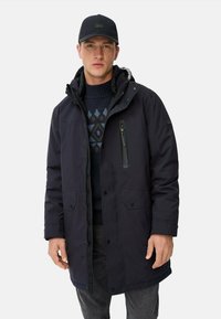 Mørkeblå polstret parka med en lynlåslukket brystlomme, trykknaplukning foran og justerbar hætte. Båret over en mønstret sweater og mørke bukser.