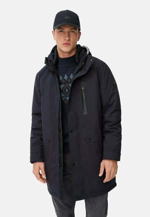 Marineblauer gepolsterter Parka mit einer mit Reißverschluss versehenen Brusttasche, Druckknopfverschluss und verstellbarem Kapuzen. Über einem gemusterten Pullover und dunklen Hosen getragen.