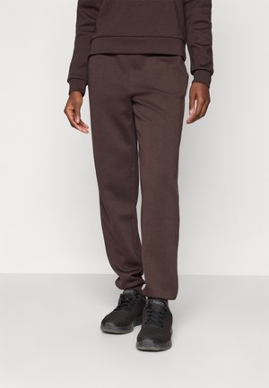 Person iført mørkebrune joggingbukser og matchende sweatshirt parret med sorte sportssko, stående mod en ensfarvet hvid baggrund.