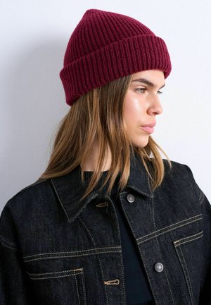 Young woman with straight brown hair wearing a maroon knit beanie and a dark denim jacket, looking to the right against a plain background.