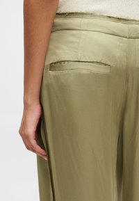 Pantalon vert olive avec une texture lisse, doté d'une poche arrière et d'une fente sur le côté. La taille présente un léger détail de surpiqûre contrasté.